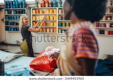Smiling printing shop worker standing at facility with color swatch in her hands and choosing bucket of paint for printing while smiling at her multicultural colleague in foreground.Women in industry.
