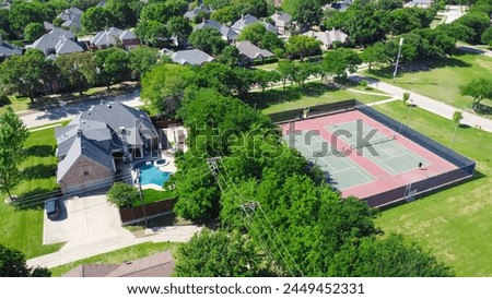 Community tennis courts with chain link fence and players in upscale residential neighborhood with large two-story single-family house, swimming pool, fenced backyard, suburb Dallas, TX, aerial. USA Royalty-Free Stock Photo #2449452331