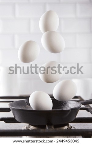 Six white eggs fall into a black cast iron pan. Kitchen scene on a gas stove. Modern and creative motif. Color mood bright, clear and fresh. Food in motion.