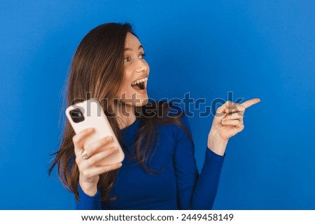 Brunette woman enjoys mobile communication and modern technologies poses against blue background points away on free space for your advertising content.