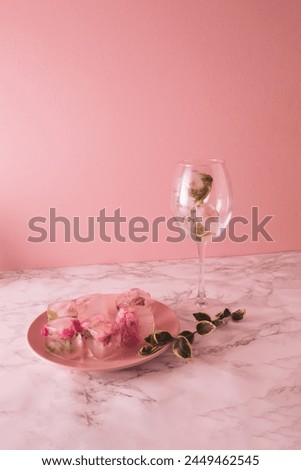 Beautiful small pink flowers in ice cubes and glass with petals on pink plate on marble and pink background.