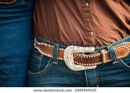 Closeup of a western style belt buckle worn by a cowgirl, she is wearing a brown shirt and blue jeans.  Royalty-Free Stock Photo #2449469635