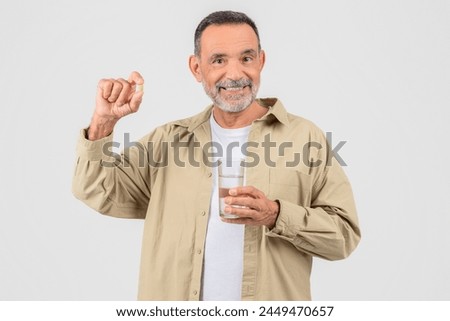 Senior man prepares to take medication, holding a pill and water, isolated on white studio background Royalty-Free Stock Photo #2449470657