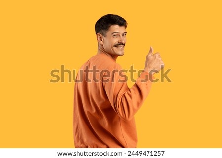 Cheerful young man with moustache in an orange sweater gives a thumbs up, isolated on a yellow background