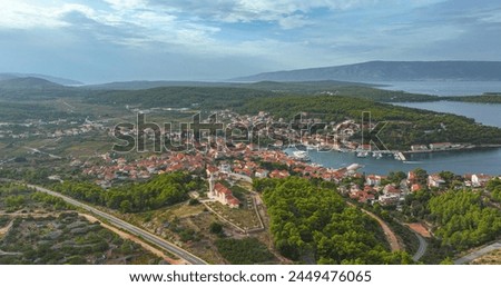 AERIAL: Flying over the lush greenery and towards the scenic marina of Hvar island, Croatia. Picturesque view of a coastal town sprawling across the hillside of a famous tourist island in the Adriatic Royalty-Free Stock Photo #2449476065