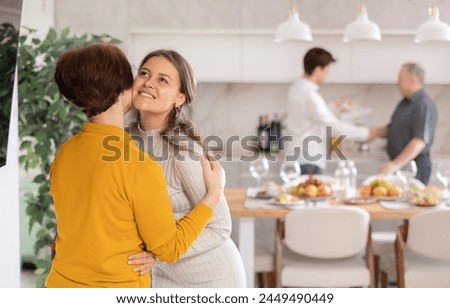 Happy young adult daughter warmly welcoming with hug and kiss senior mother coming to cozy family get-together