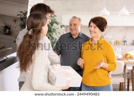 Hospitable smiling elderly couple warmly welcoming adult daughter with husband bringing gift to parents for family celebration in comfortable pleasant atmosphere at home.... Royalty-Free Stock Photo #2449490483