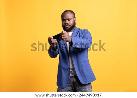 BIPOC man playing videogames on cellphone in landscape mode, enjoying leisure time. Gamer enjoying game on mobile phone, having fun, isolated over yellow studio background