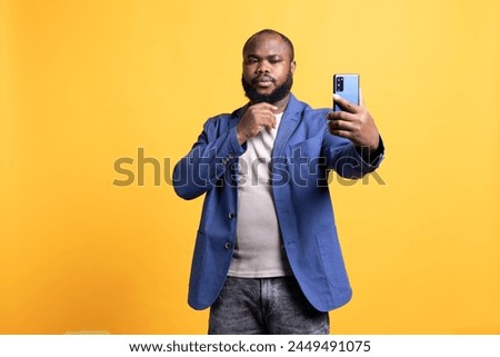 Narcissistic man using cellphone to take selfies, stroking his chin. Vain social media user taking photos using phone selfie camera, isolated over yellow studio background