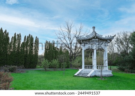A white marble Chinese garden pavilion with intricate carvings and surrounded by trees in a park. Open, airy, and octagonal design with latticework on walls and roof.