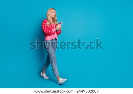 Full size photo of gorgeous person dressed knitwear jumper jeans hold smartphone walk look empty space isolated on blue color background