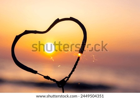 Heart-shaped sparkler burning against background of sea and rising sun at dawn. Bengal fire in shape of heart sparkling at sunrise dawn and sunset on sea. Love infatuation Valentine's Day Royalty-Free Stock Photo #2449504351