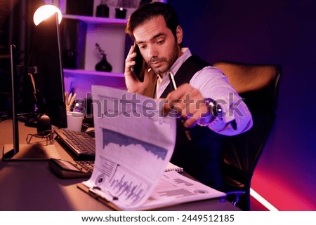 Smart businessman calling partner or investor on smartphone writing memo market data on paper work report folder to analyze sales channel discipline's product service at neon dark light room. Surmise. Royalty-Free Stock Photo #2449512185