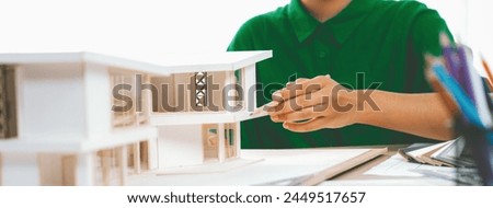 A cropped imaged of female designer measures a house model with a pencil, focusing intently on her work with house model and color swatches placed on table. Focus on hand. Closeup. Variegated.