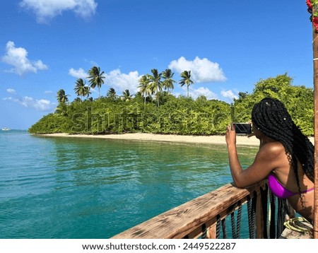 Black woman in a pink bikini leaning on a wooden rail and taking a pic on her smartphone of an island coastline with blue water, clear sky, and trees