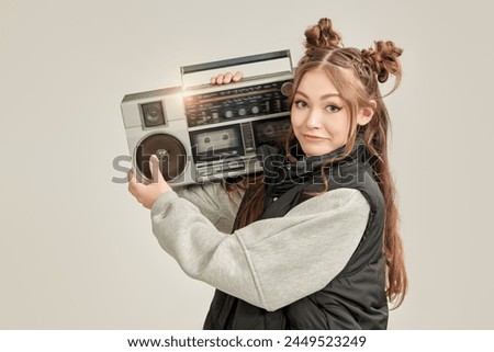 Pretty emotional teenage girl, dressed in loose casual clothes, with cute hair bumps on her head listens to music on an old boombox. Grey background with copy space. Youth style. Teens party. 