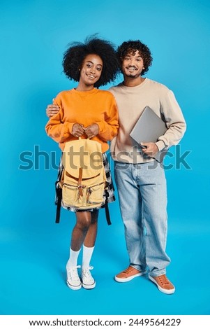 An interracial couple of students stand together in casual attire against a blue backdrop in a studio.