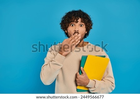 A man of Indian descent stands in casual attire against a blue backdrop, holding a folder and displaying a surprised expression.