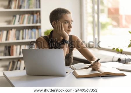 Thoughtful student girl sitting at desk with textbook and laptop, looks out window, deep in thoughts, thinking on mathematical problem, feels tired or unmotivated, disinterested in studying process Royalty-Free Stock Photo #2449569051