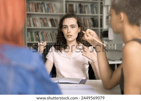 Group of schoolmates sit at desk in school library, discuss topic, share insights, ideas, opinion, focus on team leader express thoughts and solution, gathered together for studying and brainstorming Royalty-Free Stock Photo #2449569079