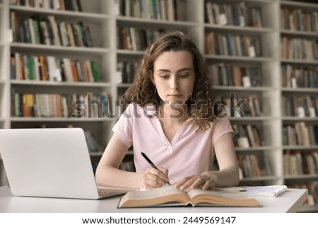 Pretty 18s student girl gain new knowledge, engaged in studying process in college library, sit at desk with laptop, read topics in textbook, makes notes, writes summary. Higher education, development Royalty-Free Stock Photo #2449569147