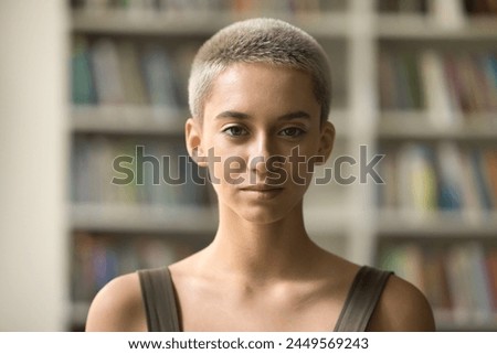 Head shot portrait serious beautiful Caucasian girl with pretty appearance and fair-haired short haircut looking at camera standing on bookshelves background. Student profile picture, study, education