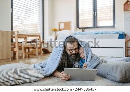 Little boy watching cartoon movie on tablet with father, lying under blanket on floor in kids room. Dad explaining technology to son, digital literacy for kids. Royalty-Free Stock Photo #2449586457