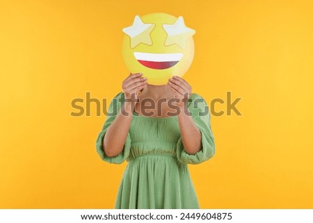 Woman holding emoticon with stars instead of eyes on yellow background