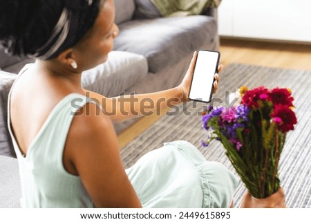African American woman holding smartphone, showing screen on a video call date with copy space. She has dark hair, wearing casual clothes, sitting with colorful flowers, unaltered Royalty-Free Stock Photo #2449615895