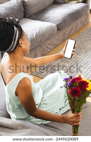 African American woman holding smartphone, showing screen on a video call date with copy space. She has dark hair, wearing a light green dress, and holding colorful flowers, unaltered.