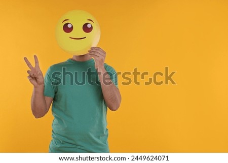 Man covering face with smiling emoticon and showing peace sign on yellow background. Space for text