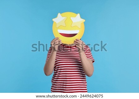 Woman holding emoticon with stars instead of eyes on light blue background