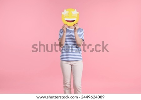 Woman holding emoticon with stars instead of eyes on pink background