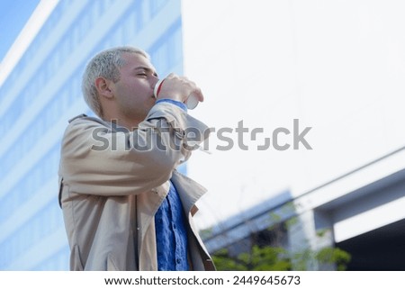 young blond Argentinian latin man, elegant with jacket and shirt, distracted outdoors standing drinking coffee and looking around with buildings in background, copy space.