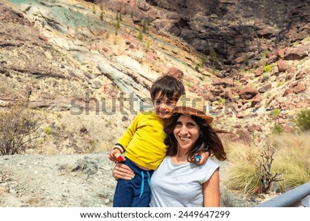A woman and a child are posing for a picture in front of a rocky mountain. The woman is wearing a straw hat and the child is wearing a yellow shirt. Scene is lighthearted and joyful, as the mother