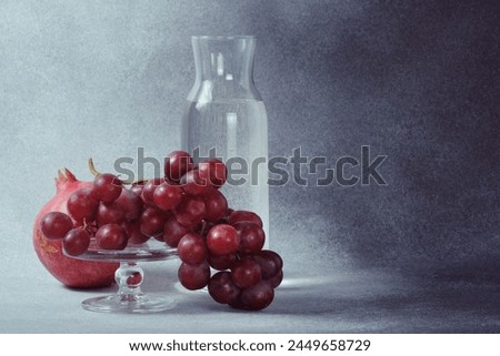 Water Carafe, Pomegranate and Red Grapes on Glass Stand, Textured Grey Background Royalty-Free Stock Photo #2449658729