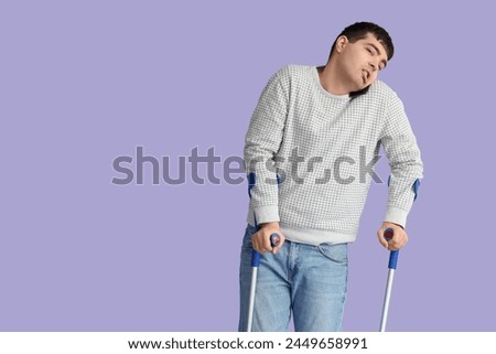 Young man with crutches talking by mobile phone on lilac background. National Cerebral Palsy Awareness Month Royalty-Free Stock Photo #2449658991