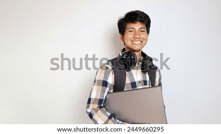 young asian male student wearing earphones and holding laptop on isolated white background
