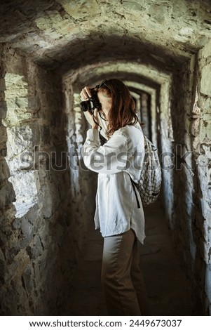 Interested female tourist takes pictures on photo camera while standing in stone corridor of ancient castle
