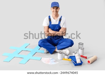 Male painter with rollers, paint cans and hashtag sign sitting on grey background