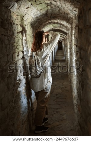 Interested female tourist takes pictures on photo camera while standing in stone corridor of ancient castle
