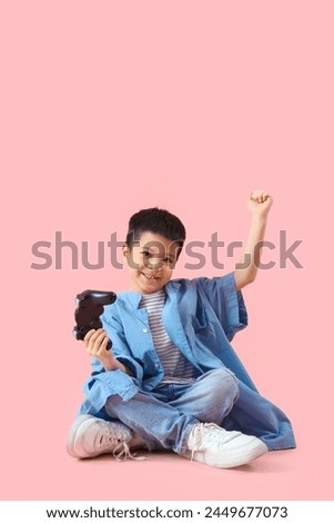 Cute little Asian boy in adult clothes with game pad on pink background