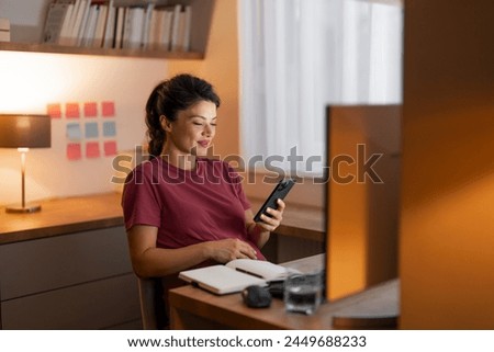 A beautiful pregnant lady using her phone to send text messages while sitting at a desk in front of a computer and working from home. Royalty-Free Stock Photo #2449688233