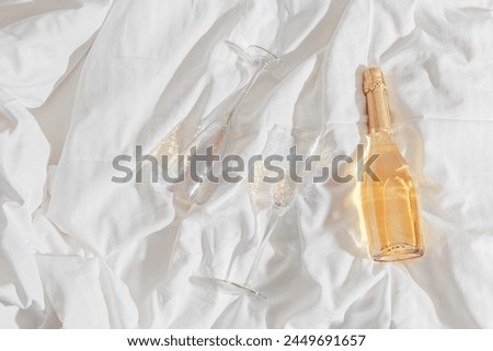 Rainbow color shining champagne glasses and white sparkling wine bottle on bed, on white blanket background. Lifestyle aesthetic photo, star filter effect. Romance meeting, romantic holiday concept. 