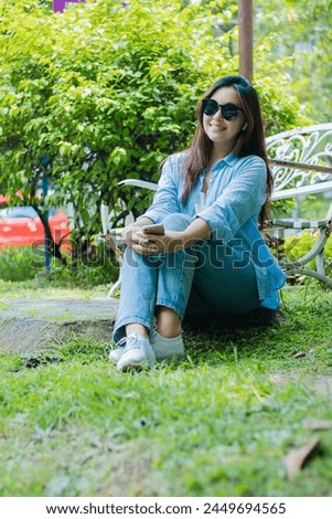 Asian mature girl wearing sunglasses smiling at the camera carrying a cell phone gadget sitting on the grass in an outdoor park. female students for the themes of education, technology and lifestyle