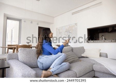 Positive young Indian homeowner girl turning ventilation cooling system on, holding remote control device, starting air conditioner at home, sitting on couch in cozy apartment