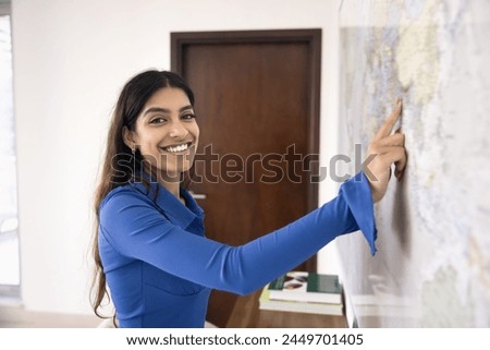 Happy young Indian woman pointing finger at world geography map on wall, looking at camera for portrait with toothy smile, planning travel, vacation, enjoying future foreign trip