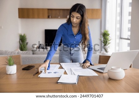 Serious young Indian startup manager girl reviewing project documents, marketing reports at home office workplace table, doing paperwork, working on marketing strategy, business plan
