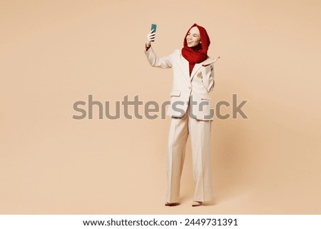 Full body young Arabian Asian Muslim woman wears red abaya hijab suit clothes doing selfie shot on mobile cell phone waving hand isolated on plain beige background. UAE middle eastern Islam concept