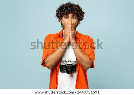 Traveler shocked Indian man wear orange casual clothes cover mouth with hand isolated on plain blue background. Tourist travel abroad in free spare time rest getaway. Air flight trip journey concept
